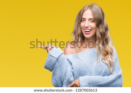 Beautiful young blonde woman wearing winter sweater over isolated background with a big smile on face, pointing with hand and finger to the side looking at the camera.