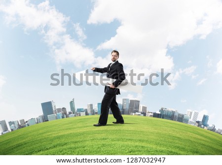 Confident and young businessman in suit holding big white arrow in hands which pointing to the side while standing on green lawn and cityscape view on background.