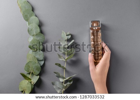 Female hand with bottle of perfume and eucalyptus branches on grey background