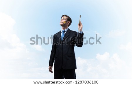 Horizontal shot of young and happy businessman in black suit holding paintbrush in his hand while standing against blue and cloudy skyscape on background.