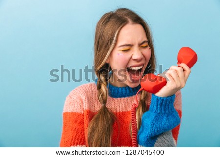 Excited young girl wearing winter clothes standing isolated over blue background, talking on a landline phone