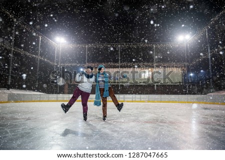 two cute girls skating and having fun on outdoor open air ice-rink at winter at night while snow falling