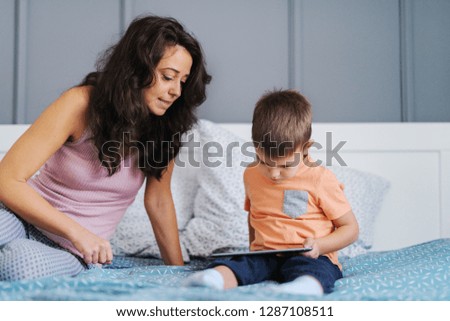 Little kid watching cartoons on tablet while sitting on the bed while his mother watching cartoons with him. Bedroom interior.