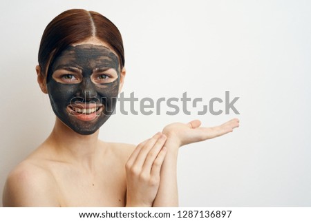Joyful woman in a clay cosmetic mask holds a free space on her hand on a light background