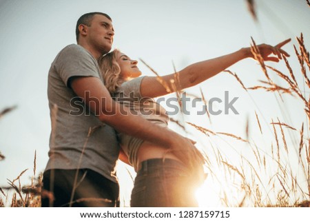 A young man embraces the belly of his pregnant wife during a walk against background of sky and spikelets. Backlight.