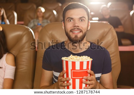 Excited male viewer keeping popcorn in hands and watching interesting film in cinema. Young bearded man wearing shirt expecting final of movie. Concept of entertainment and leisure. Royalty-Free Stock Photo #1287178744