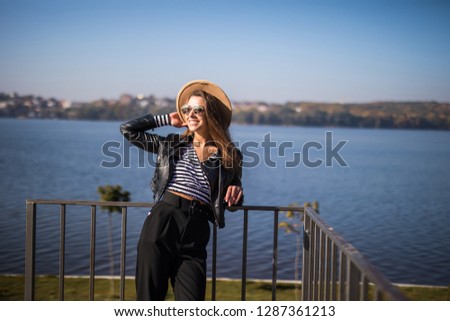 Young woman with fanky hat and sunglasses standing on city pier on promenade of park lake