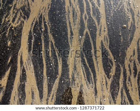 Close up of a natural sand pattern where dark silt is cut through by the receding tide to reveal a lighter yellow sand. Small grit, stones and shells are left behind. A vertically braided natural art.