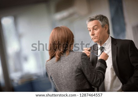 Businessman rests the end of a temple arm of a set of eyeglasses in the corner of his mouth and raises his eyebrows in surprise as he listens to a female colleague.