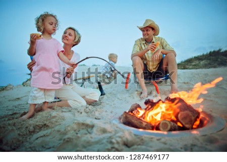 Happy young family have fun cooking sausages for their dinner over a glowing campfire on the beach at twilight.