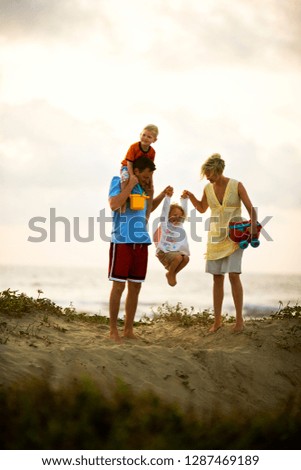 Family at the beach at sunset swing their daughter in the air while their son sits on his daddy's shoulders.