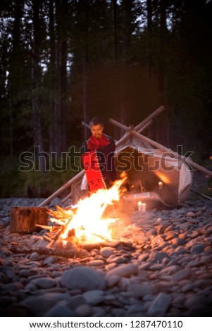 Mid-adult man with a blanket around him as he stands beside a bonfire on a rocky beach