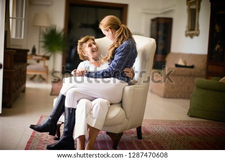Mid-adult woman sitting on her mature mother's knee in a living room.