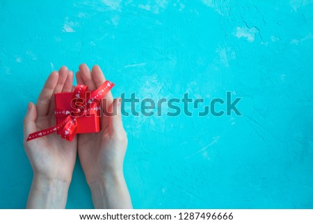 Female hands hold a red gift box with a bow on blue background. Happy Valentine’s Day card.