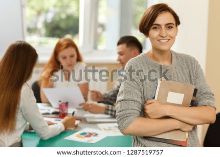Portrait of young female student keeping book and notes, looking at camera, smiling and posing during lesson on courses. Students studying and talking at background. Interesting learning concept.