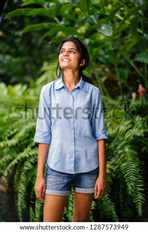 A young Indian Asian girl stands in a park amidst lush green plants during a warm summer day. She is wearing a casual blue shirt and denim shorts. and is smiling happily. 