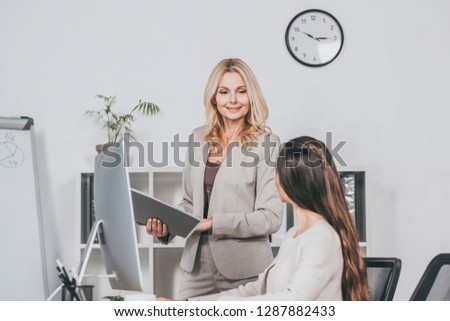 smiling mature businesswoman holding folder and looking at young colleague working with desktop computer in office