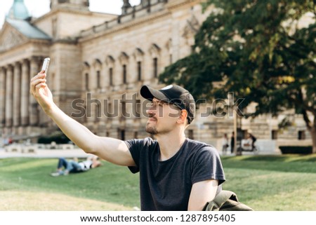 A young male tourist or student sits on the grass outside in Leipzig, Germany, talking on video or using a mobile phone to chat or take pictures.
