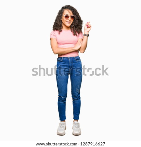 Young beautiful woman with curly hair wearing pink sunglasses with a big smile on face, pointing with hand and finger to the side looking at the camera.