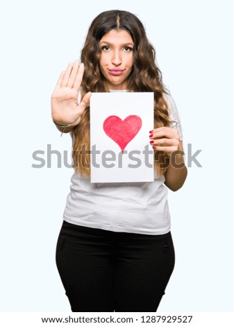 Young adult woman holding card with red heart with open hand doing stop sign with serious and confident expression, defense gesture