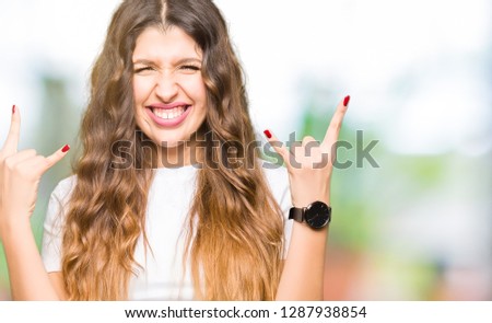 Young beautiful woman wearing casual white t-shirt shouting with crazy expression doing rock symbol with hands up. Music star. Heavy concept.