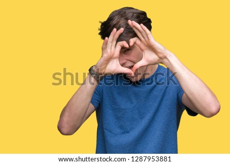 Young handsome man wearing blue t-shirt over isolated background Doing heart shape with hand and fingers smiling looking through sign