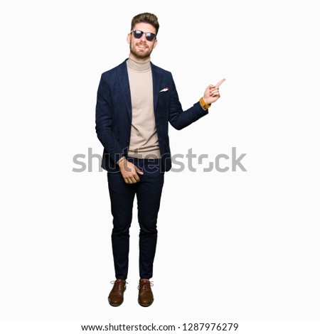 Young handsome elegant man wearing sunglasses with a big smile on face, pointing with hand and finger to the side looking at the camera.