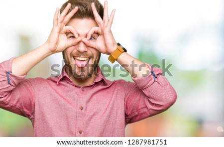 Young handsome business casual man doing ok gesture like binoculars sticking tongue out, eyes looking through fingers. Crazy expression.