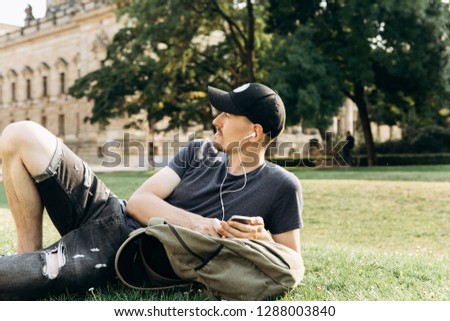 A young guy with a backpack or a student lies on the grass and listens to music or a podcast and enjoys.