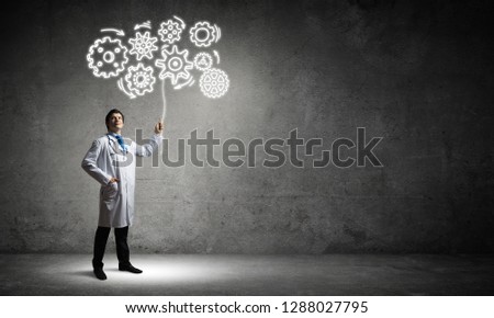 Conceptual image of young and successful doctor in white uniform interracting with gear mechanism structure while standing against gray wall on background.