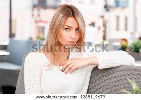 Cute attractive young woman in a beautiful smile with blue eyes with blond hair in a stylish white knitted sweater sits in an outdoor cafe. Charming girl on vacation.