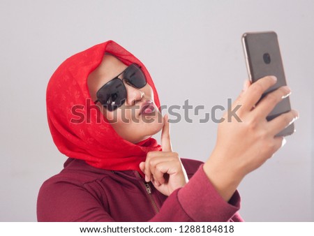 Portrait of muslim lady wearing black sunglasses in red suit and hijab smiling and taking selfie picture photograph of her self on smart phone