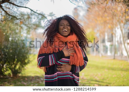 Joyful African American student keeps hands crossed, laughs at good joke, wears casual clothes and round spectacles, standing in a park. Happy young woman with curly hair poses outdoors. Imagen.
