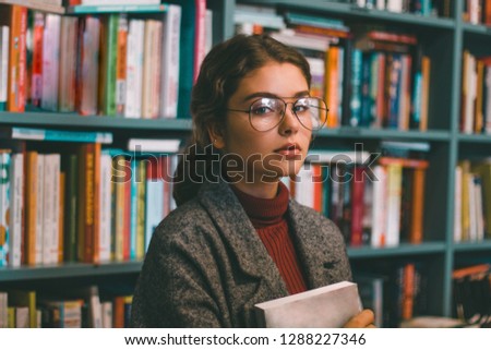 Portrait of a beautiful, intelligent - looking student reading book in library leaning against bookshelves at the university.Knowledge concept. Girl with book school building background.Talented cute 