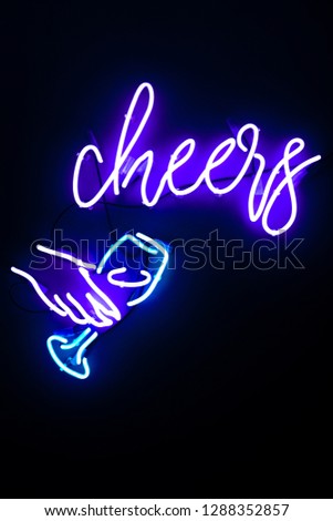 Close up neon sign design element on wall with lettering font at wine bar of blue and purple colour on dark background. Female hand holding glass of wine. cheers text on the wall. Neon tube, led wall