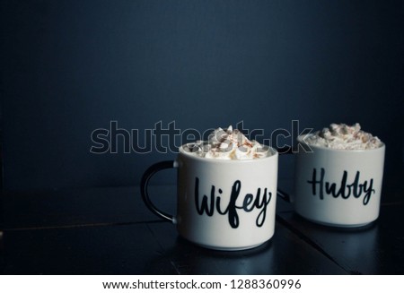 Two white coffee mugs with black handles that say wifey and hubby with whipped cream lattes in them on a dark stained wood table with a black background. Royalty-Free Stock Photo #1288360996