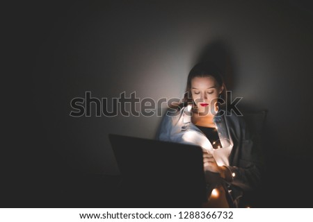 Attractive girl uses a laptop at night. Young beautiful woman lying on bed in cozy room working on laptop. Laptop on the knees of an attractive woman at night. Deadline