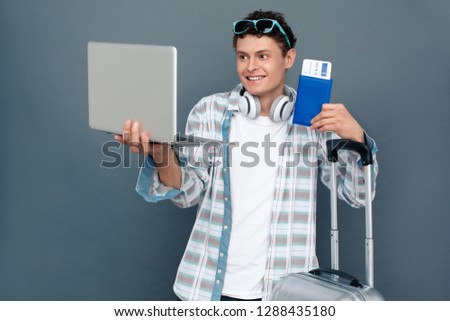Young guy wearing sunglasses and headphones isolated on gray wall tourism concept standing with luggage suitcase holding laptop having online video call showing passport with plane ticket looking