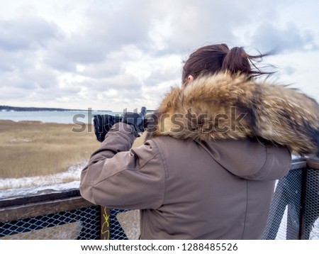 Young  tourist girl take Pictures by smartphone overlooking Baltic sea, Back view. vacation concept. Copy space. Gray sky and view from the viewing tower. Winter seascape and yellow grass