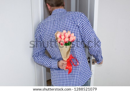 A young man in a blue plaid shirt and jeans, holding a bouquet of tulips behind his back, and peeks in the open door, for white background. Valentine's Day and March 8 concept.