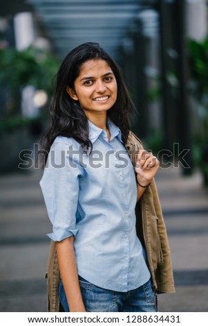 Portrait of a young and beautiful Indian Asian girl exploring the city on her own. She is casually but stylishly dressed and has a brown jacket thrown over her shoulder.