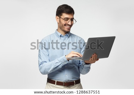 Portrait of young handsome male teacher in blue shirt holding laptop and smiling, isolated on gray background
