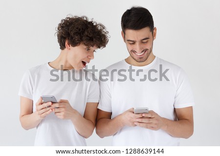 Surprised young woman looking at smartphone screen of his boyfriend with mouth open, isolated on gray background