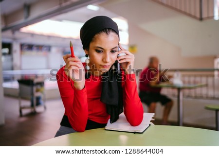 A young Muslim woman business entrepreneur is smiling as she talks on her smartphone and takes notes for her business during the day. She is wearing a hijab head scarf and smiling. 