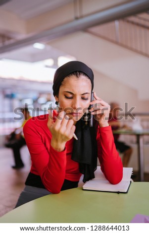 A young Muslim woman business entrepreneur is smiling as she talks on her smartphone and takes notes for her business during the day. She is wearing a hijab head scarf and smiling. 