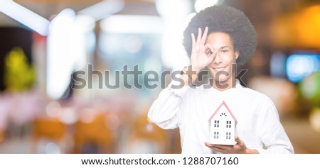 Young african american man with afro hair holding house with happy face smiling doing ok sign with hand on eye looking through fingers