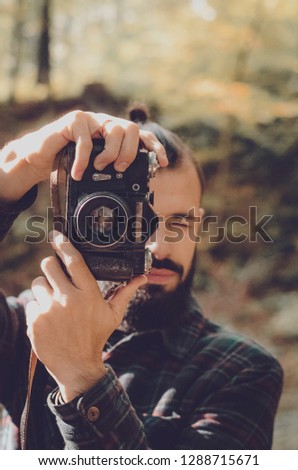 Nature Photography Concepts Professional photographer: stylish bearded man, dressed in a shirt and with a hairdo Top Knot with retro photo camera in hands takes pictures in the woods