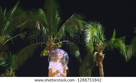 happy teen girl holding big airy, glowing balloon in her hands against the background of palm trees. evening time. concept of tourism and beach holiday.