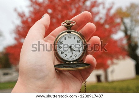 A hand holding a pocket watch. A red tree can be seen in the background out of focus. Time reads 4:27.