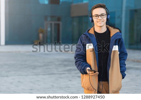Smiling young guy listening to music, holding the phone outdoors. Copy space